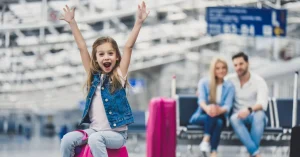 Excited young girl at an airport with parents in the background and luggage nearby.